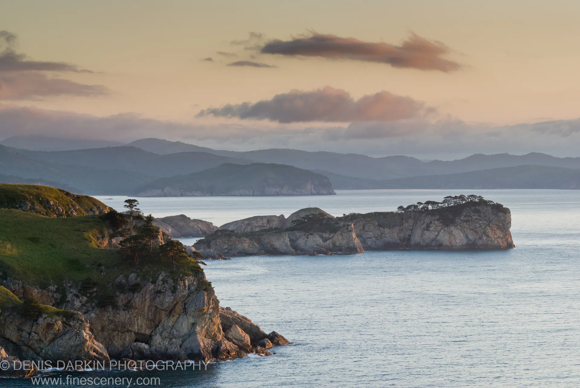 Rugged coastal cliffs in early dawn light at Gamov Peninsula, mist-covered hills receding in layers above a deep blue bay under a peach-tinted sky.