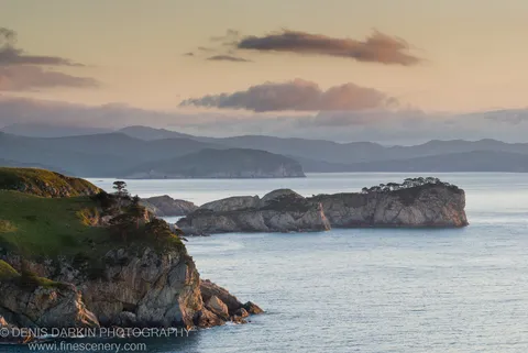 Rugged coastal cliffs in early dawn light at Gamov Peninsula, mist-covered hills receding in layers above a deep blue bay under a peach-tinted sky.