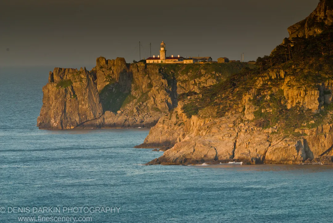 A lighthouse anchors the ridge above stratified cliffs at Gamov Peninsula, warm dawn light on the rock face and deep blue water below.