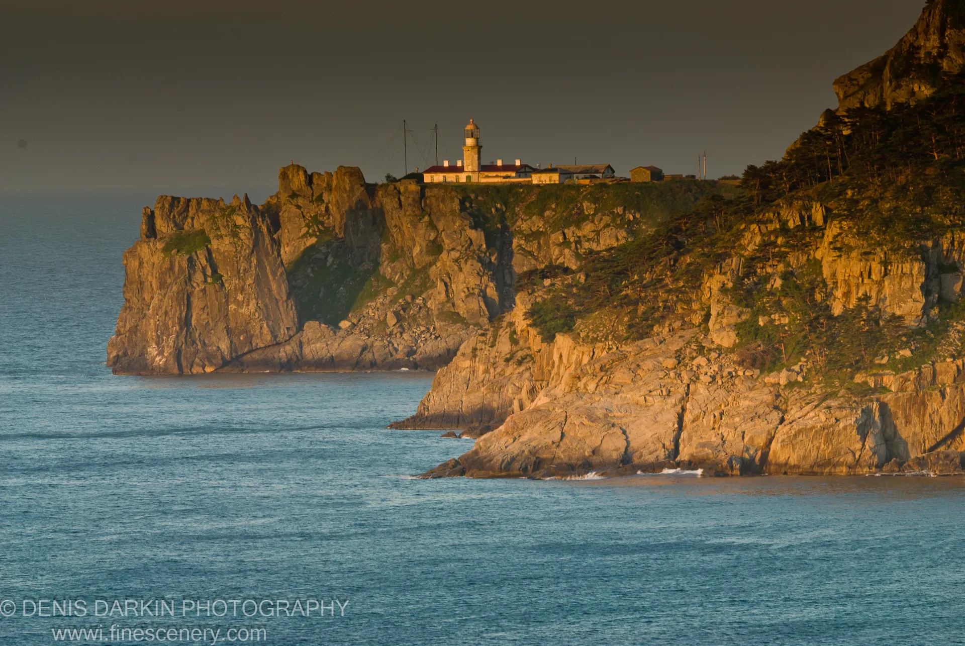 A lighthouse anchors the ridge above stratified cliffs at Gamov Peninsula, warm dawn light on the rock face and deep blue water below.