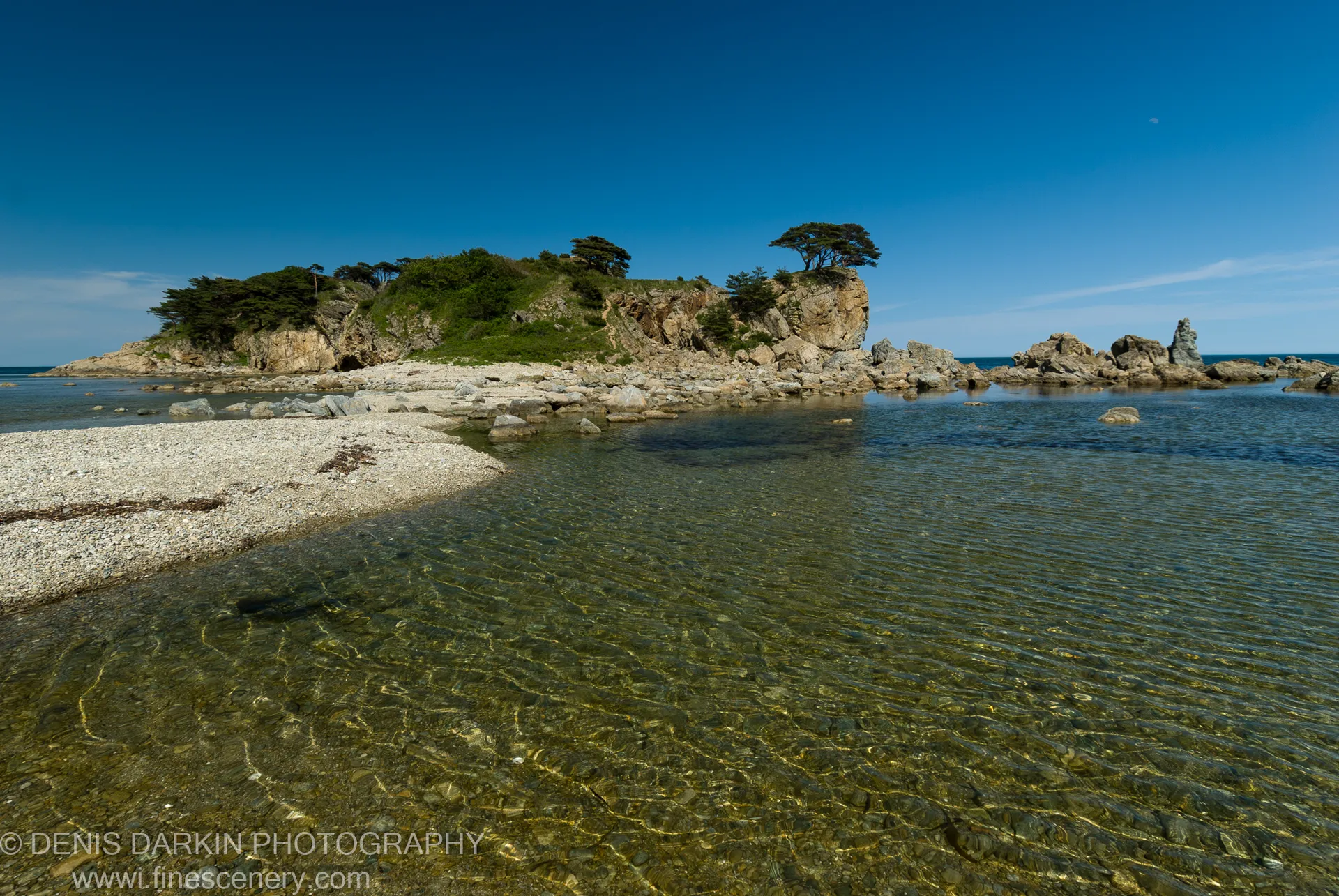 Clear rippling shallows at Gamov Peninsula reveal a sandy bottom, a grassy headland with dark evergreen trees rising above the rocks beyond.