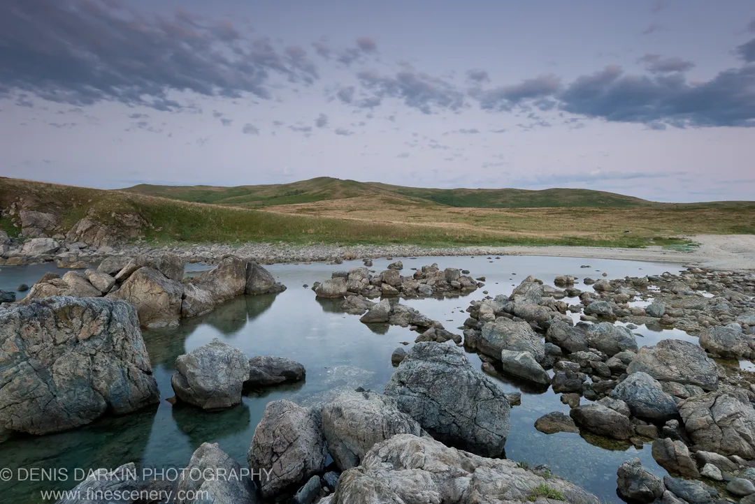 Weathered boulders emerge from glass-smooth water at Gamov Peninsula at dawn, rolling hills of grass rising beyond under a pale sky.