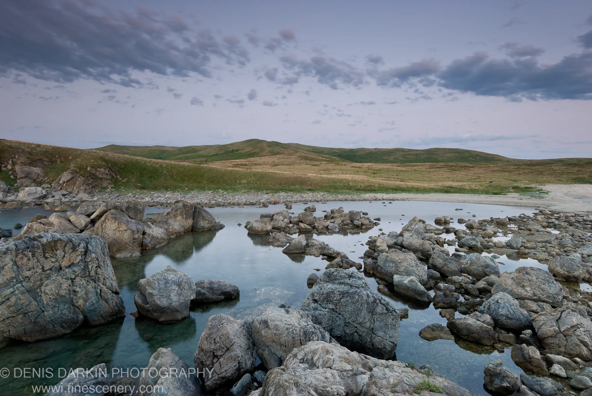 Weathered boulders emerge from glass-smooth water at Gamov Peninsula at dawn, rolling hills of grass rising beyond under a pale sky.