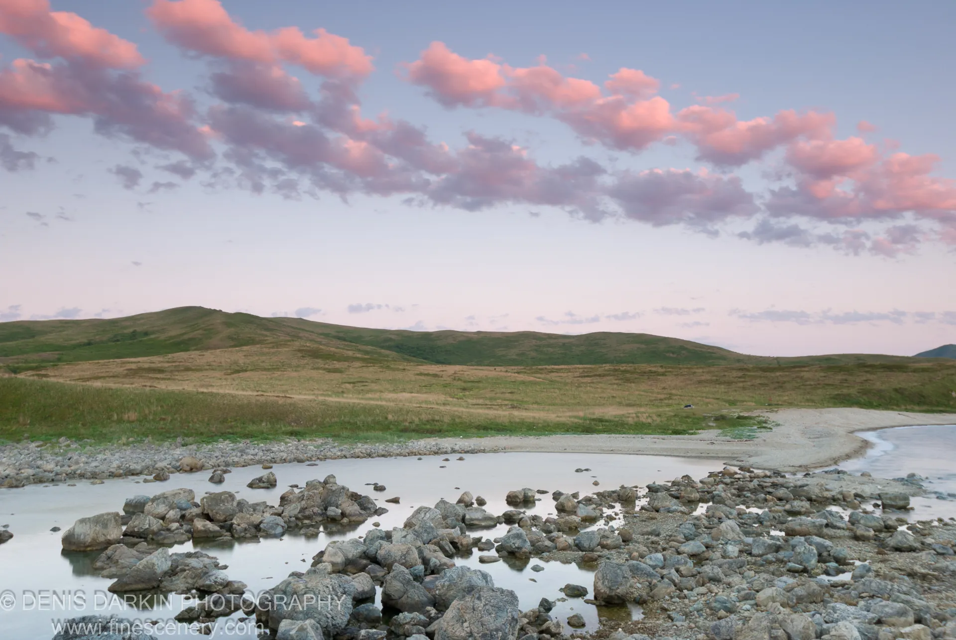 Soft pink clouds above rolling green hills at sunrise, scattered grey stones across the shallow water of a rocky shore at Gamov Peninsula.