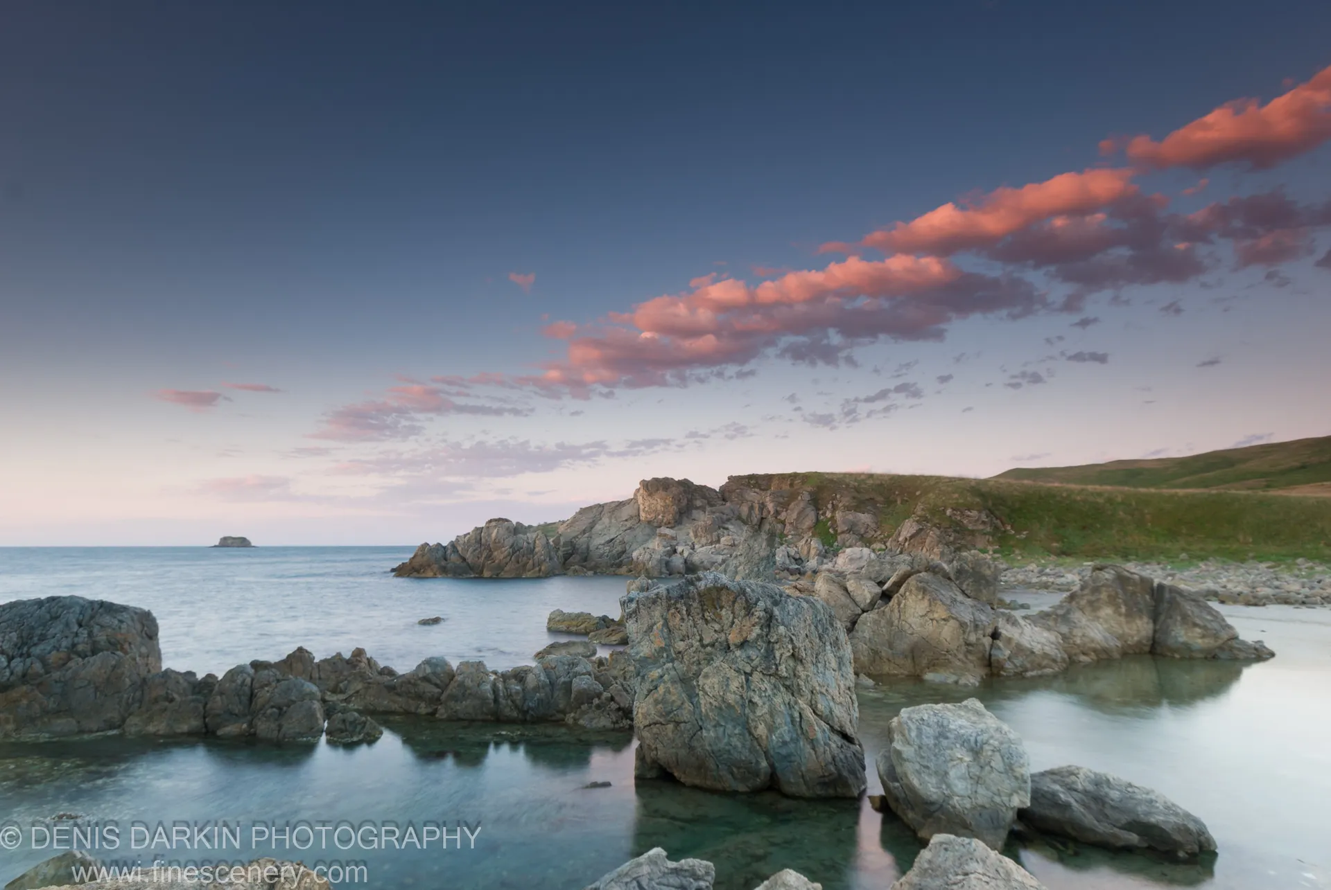 Glass-smooth water and boulders at Gamov Peninsula at dawn, a grassy headland on the right and a distant rock formation on the horizon.