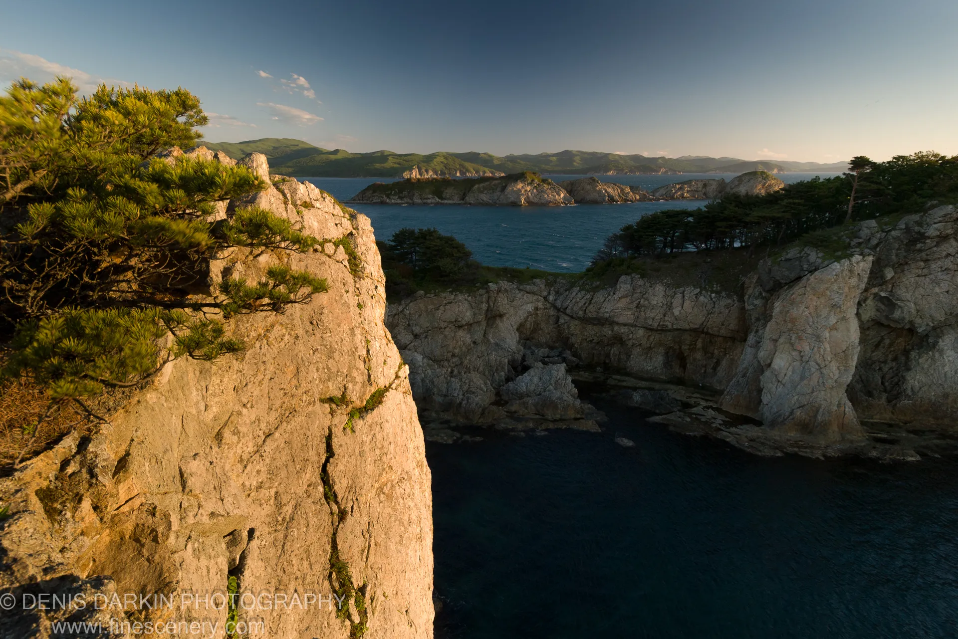 Warm sunlight on a textured cliff face in the foreground, with a deep blue cove and rolling hills visible below and beyond, Vityaz Bay.