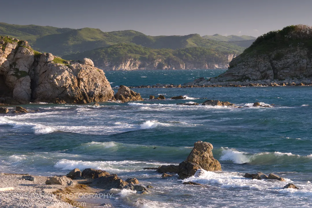 White-capped waves churning around boulders on a pebble beach below rugged cliffs, with a blue-green bay and rolling hills beyond, Gamov Peninsula.