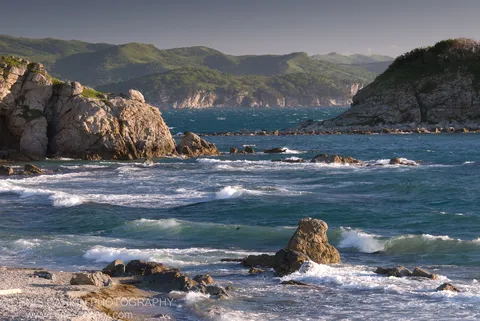 White-capped waves churning around boulders on a pebble beach below rugged cliffs, with a blue-green bay and rolling hills beyond, Gamov Peninsula.