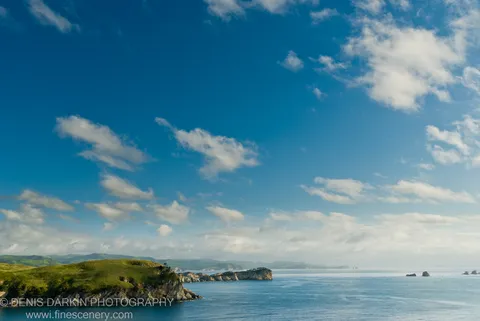 Lush green coastal hills slope to calm blue water at Gamov Peninsula, with sea stacks and cumulus clouds filling an azure sky overhead.