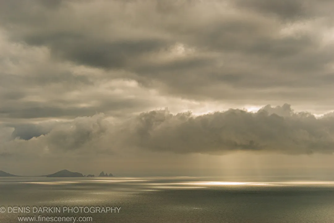 A shaft of sunlight breaks through thick storm clouds above Vityaz Bay, casting a bright reflection across the flat calm sea, with silhouetted distant islands on the horizon.