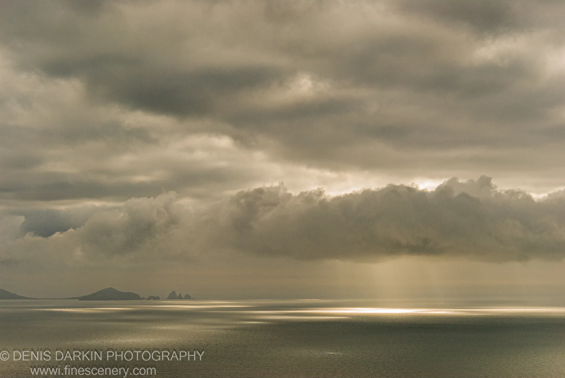 A shaft of sunlight breaks through thick storm clouds above Vityaz Bay, casting a bright reflection across the flat calm sea, with silhouetted distant islands on the horizon.