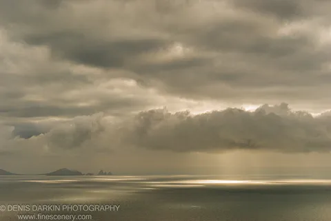 A shaft of sunlight breaks through thick storm clouds above Vityaz Bay, casting a bright reflection across the flat calm sea, with silhouetted distant islands on the horizon.