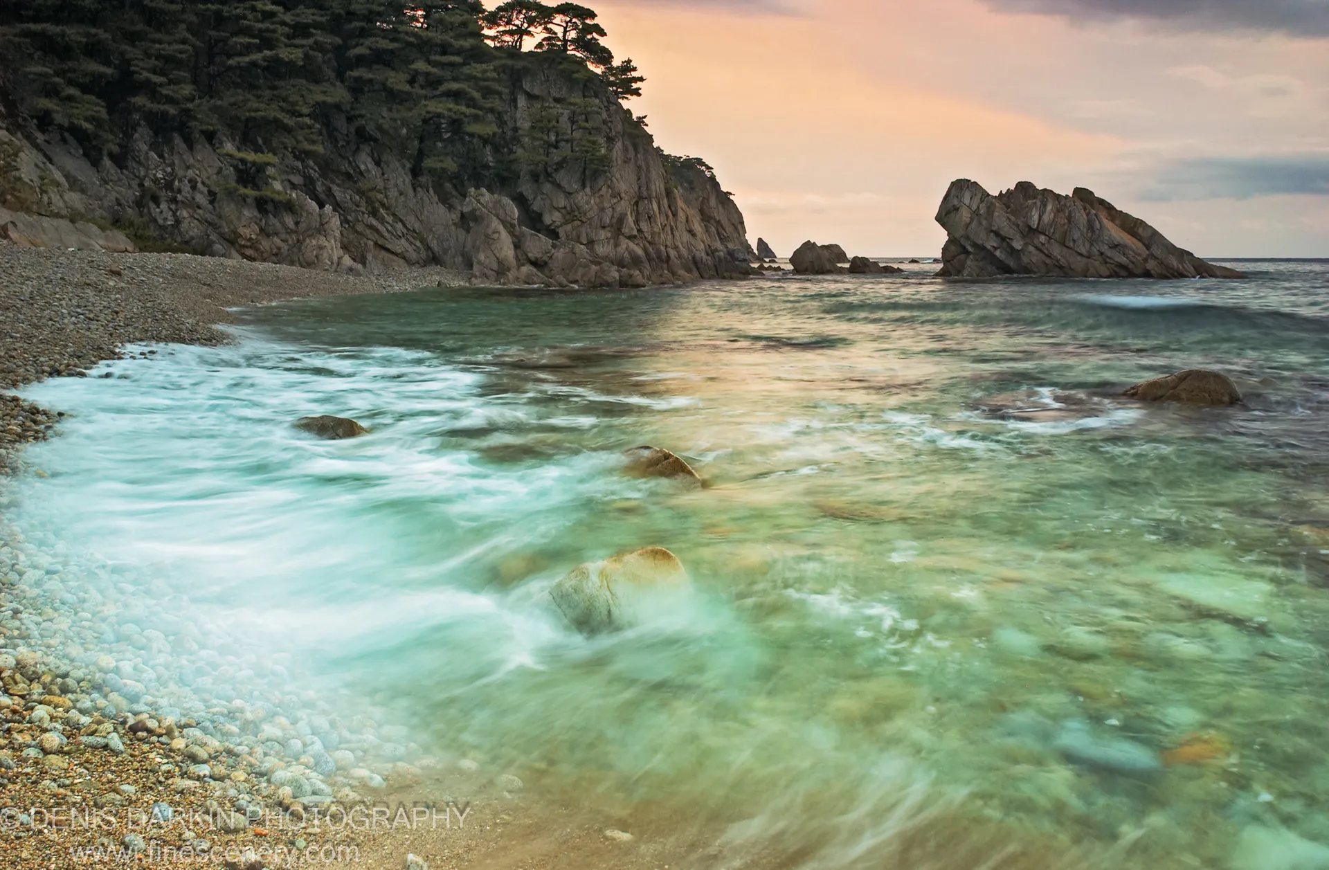 A steep pine-covered cliff rises above smooth pebbles on the Vityaz Bay shoreline, with a solitary rock formation visible at sea.