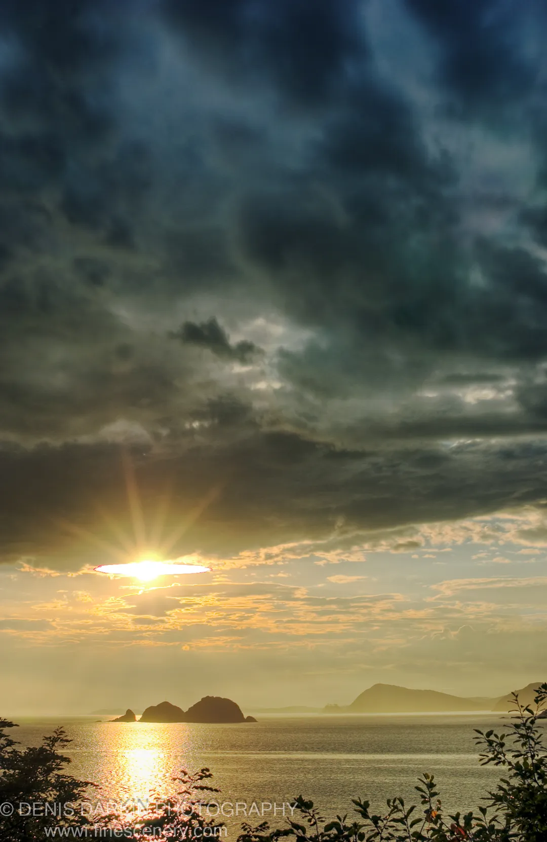 Heavy storm clouds over Vityaz Bay with a sunburst breaking through on the left, casting reflected light across the calm sea toward silhouetted rocky islands.