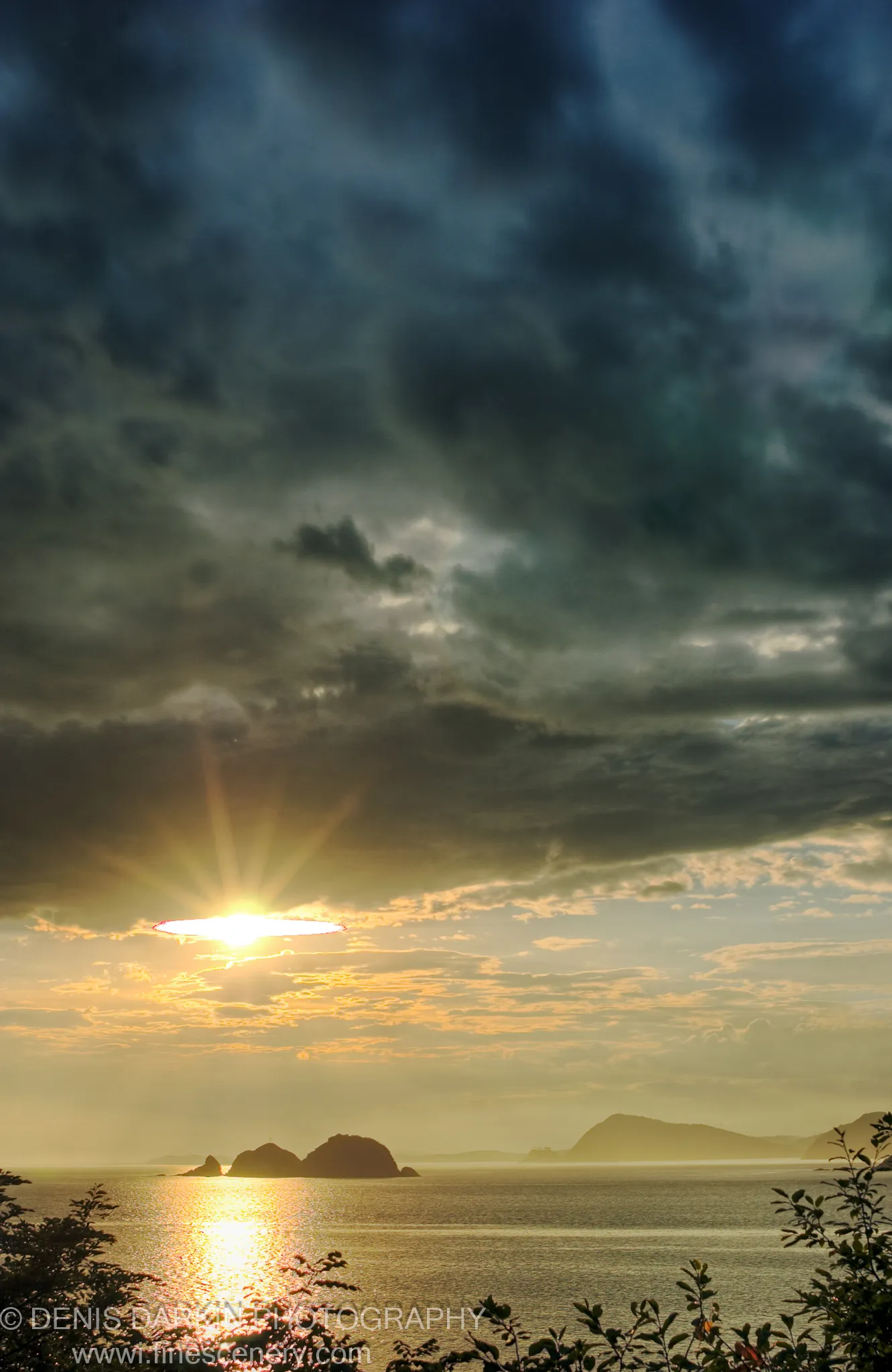 Heavy storm clouds over Vityaz Bay with a sunburst breaking through on the left, casting reflected light across the calm sea toward silhouetted rocky islands.