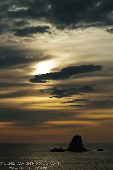 Silhouetted sea stacks rise from calm water at Gamov Peninsula as a morning sunburst breaks through a band of dark clouds in early autumn.