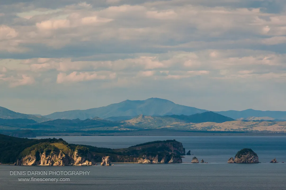 Layered mountain ridgelines recede into atmospheric haze above the Far East Marine Reserve coastline under a soft cloud-streaked sky.