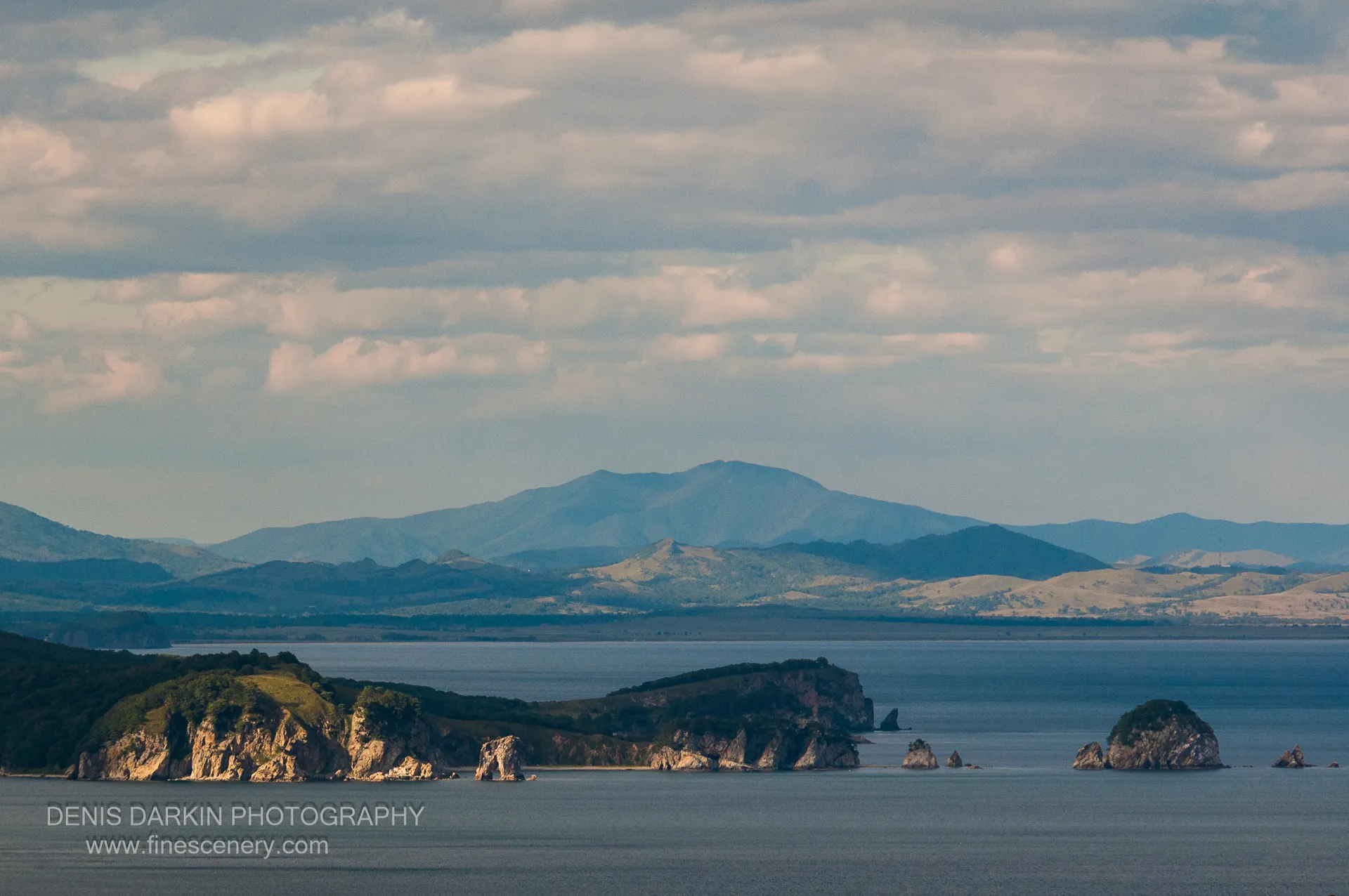 Layered mountain ridgelines recede into atmospheric haze above the Far East Marine Reserve coastline under a soft cloud-streaked sky.