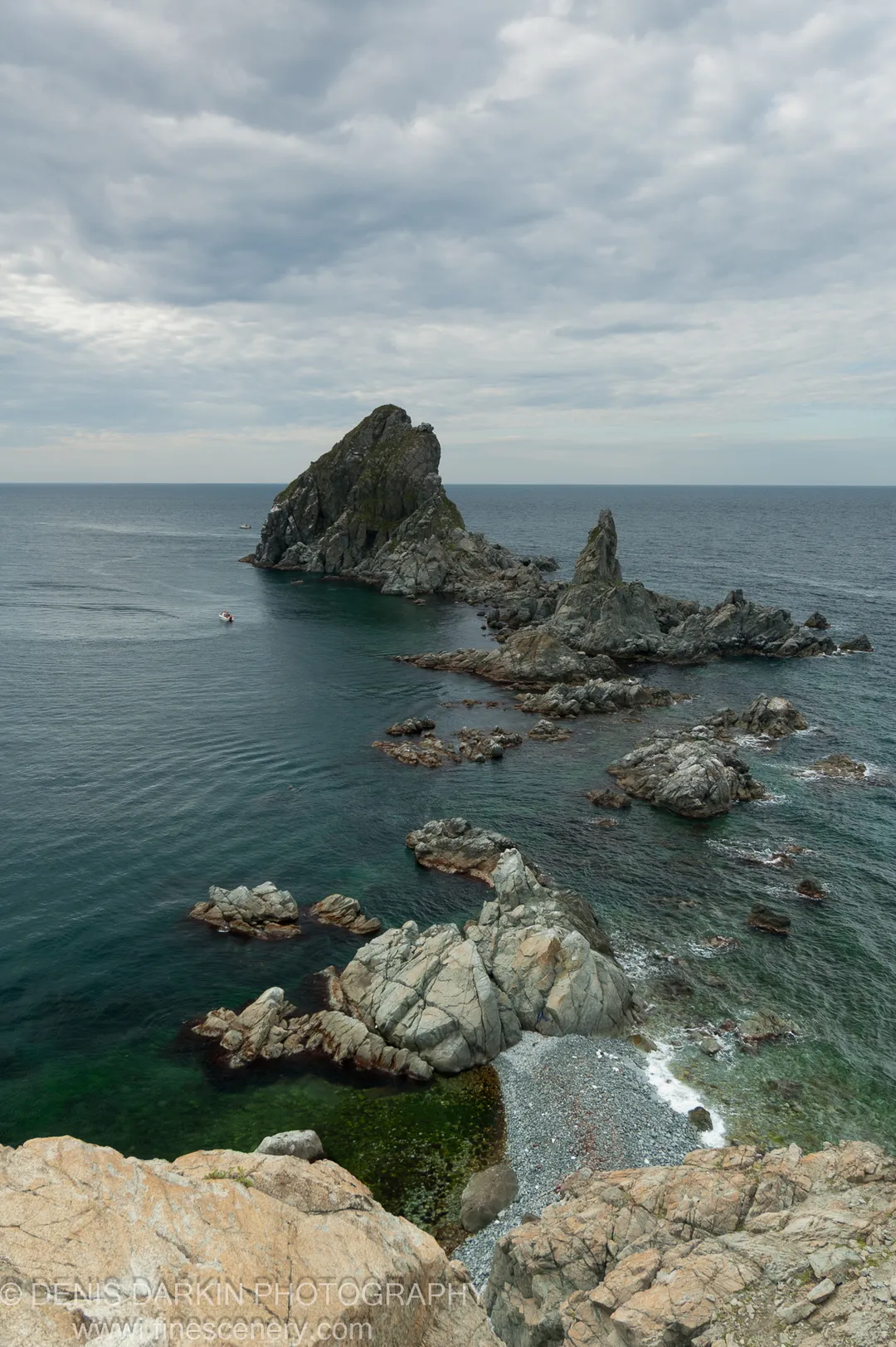 Turquoise shallows deepen to navy at Gamov Peninsula, with a rocky islet and small boat visible under an overcast midday sky.