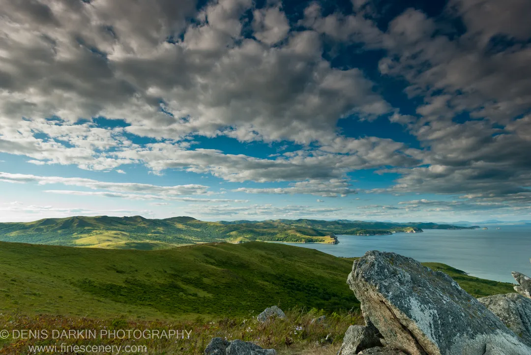 Weathered granite boulders on a Gamov Cape hilltop, with grassy slopes descending to a deep blue bay and textured cumulus clouds above.