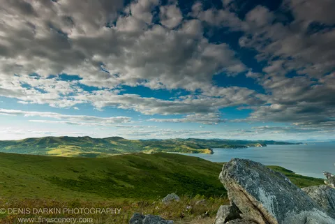 Weathered granite boulders on a Gamov Cape hilltop, with grassy slopes descending to a deep blue bay and textured cumulus clouds above.