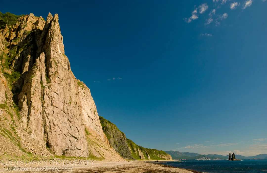 Stratified pale cliff face rising from a pebble beach at Rudnaya Pristan, with a solitary sea stack visible in the distance against blue sky.