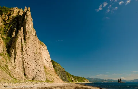 Stratified pale cliff face rising from a pebble beach at Rudnaya Pristan, with a solitary sea stack visible in the distance against blue sky.