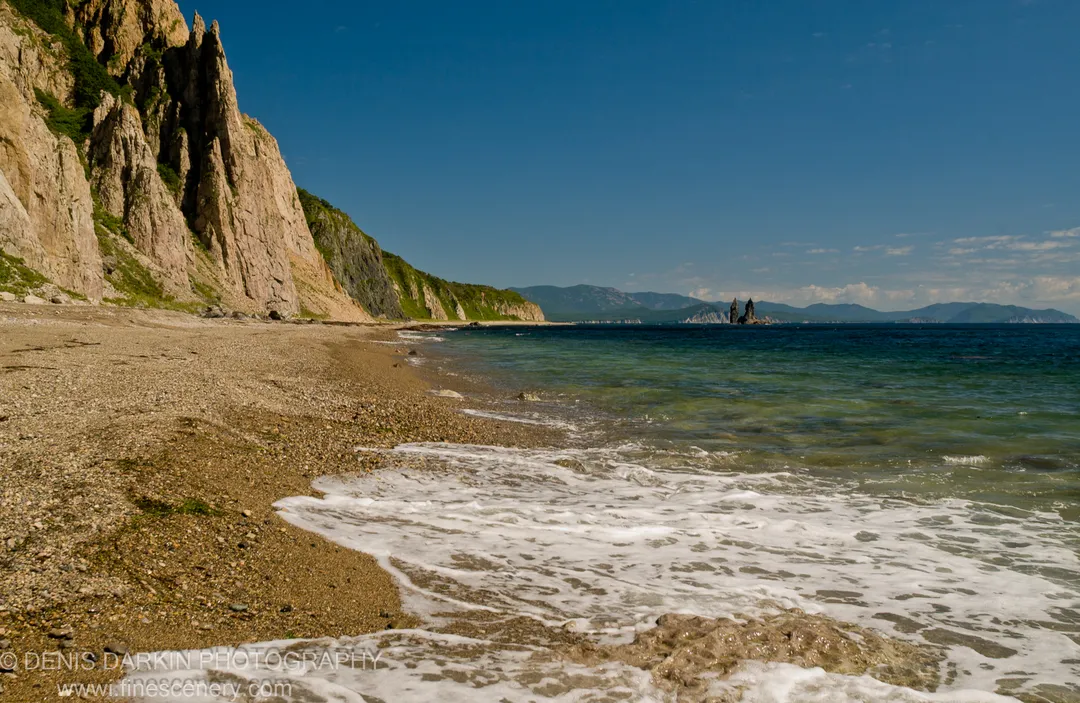 Coarse pebble beach with foamy surf and sheer tan cliffs along the shoreline at Rudnaya Pristan, Primorsky Krai, summer morning.