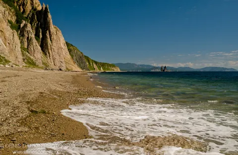Coarse pebble beach with foamy surf and sheer tan cliffs along the shoreline at Rudnaya Pristan, Primorsky Krai, summer morning.