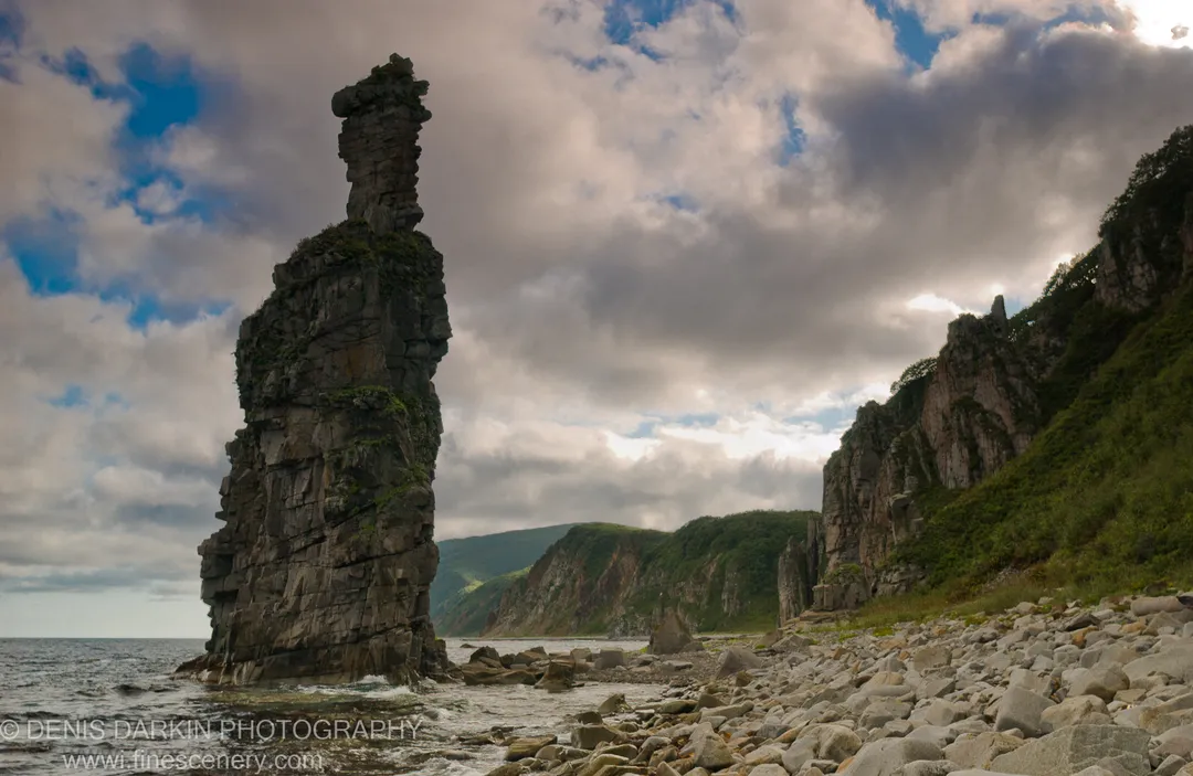 A tall sea stack rises from the pebble shore at Dalnegorsk, with steep vegetated cliffs and rolling hills receding under a cloudy sky.