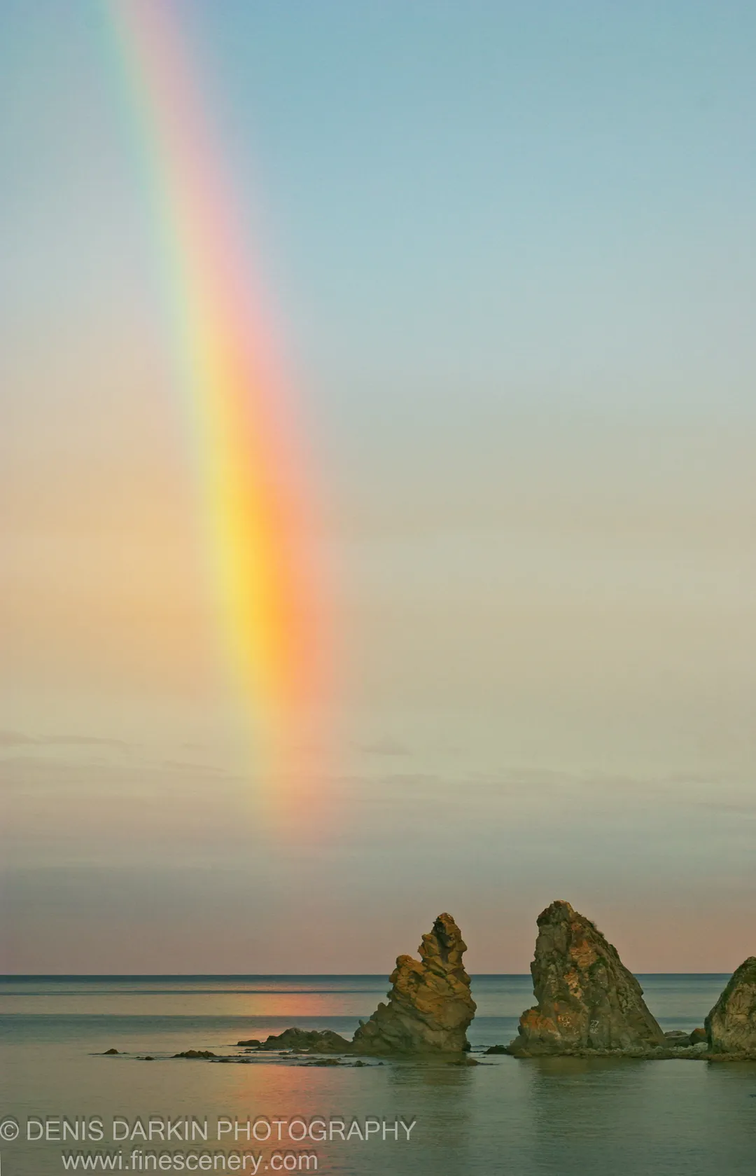 A vertical rainbow descends into a calm bay at sunset, sea stacks in the foreground against a sky of pale blue to warm peach.