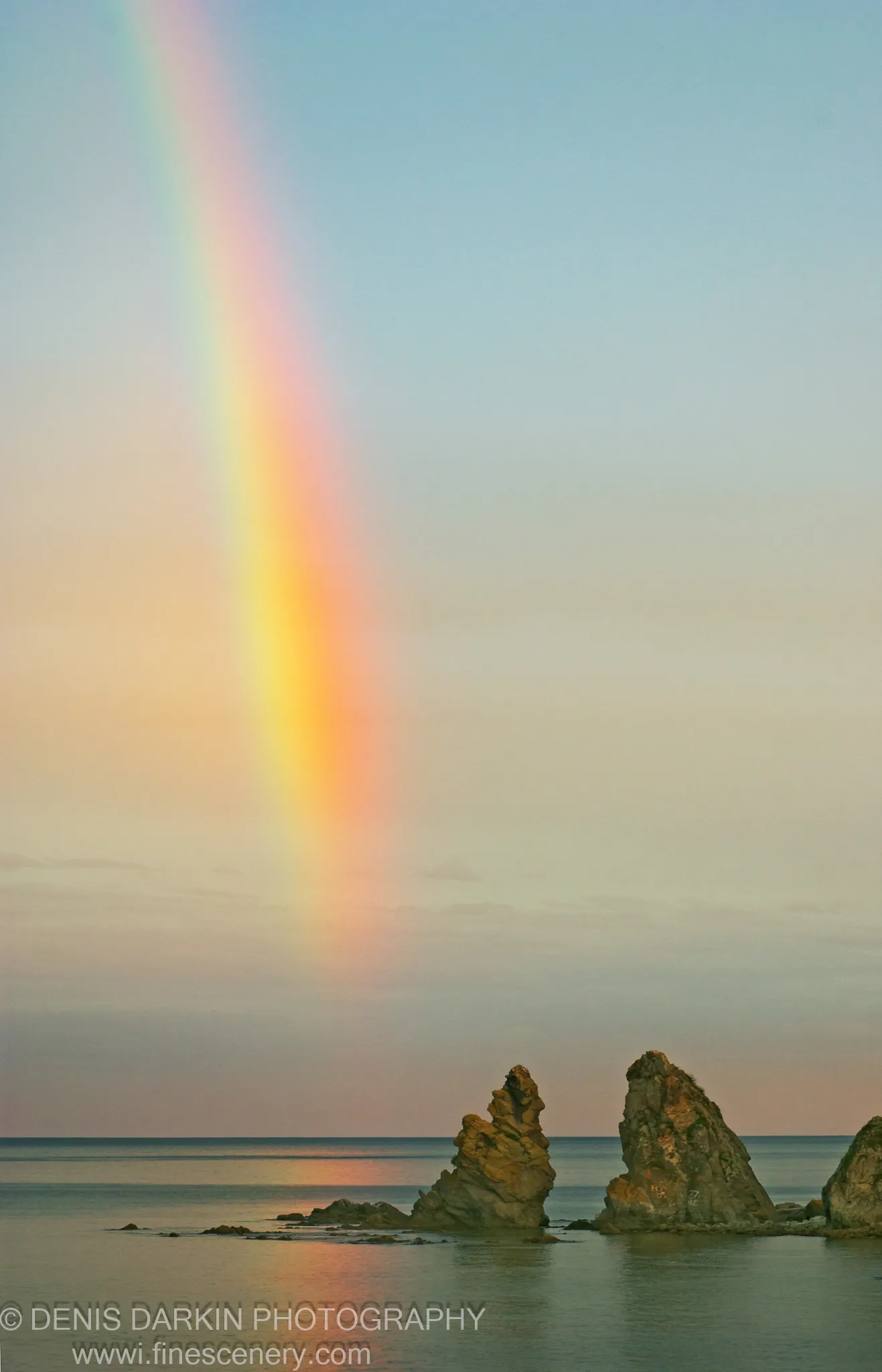 A vertical rainbow descends into a calm bay at sunset, sea stacks in the foreground against a sky of pale blue to warm peach.