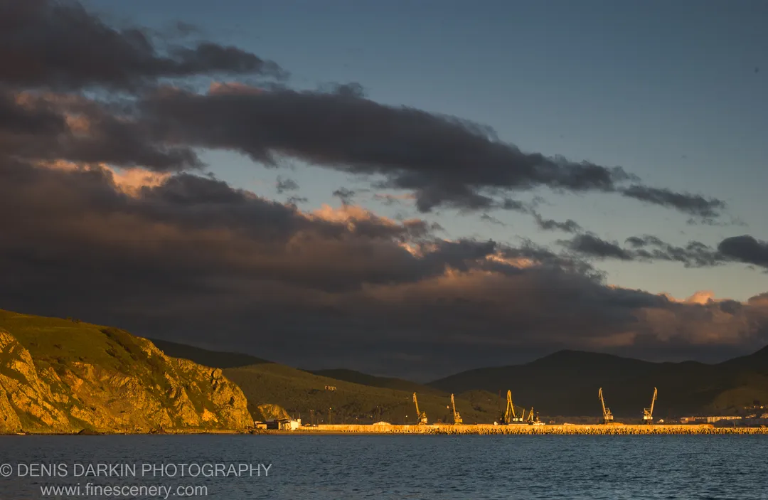 Dawn light over a harbor breakwater lined with yellow cranes, seen from a grassy cliff above the calm sea, Plastun, Primorsky Krai.