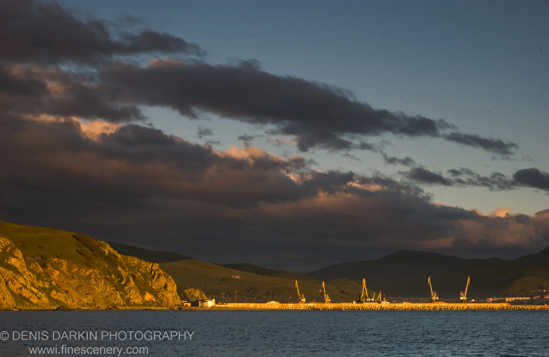 Dawn light over a harbor breakwater lined with yellow cranes, seen from a grassy cliff above the calm sea, Plastun, Primorsky Krai.