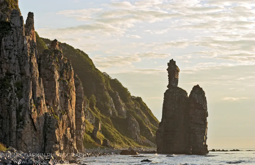 Warm dawn light on a sheer cliff face beside a sea stack in a calm bay, with soft clouds above the rocky shore.