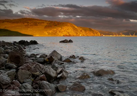 Foreground boulders beside a calm bay, a sun-lit hill beyond and industrial cranes in silhouette on the far shore, Primorsky Krai coast.