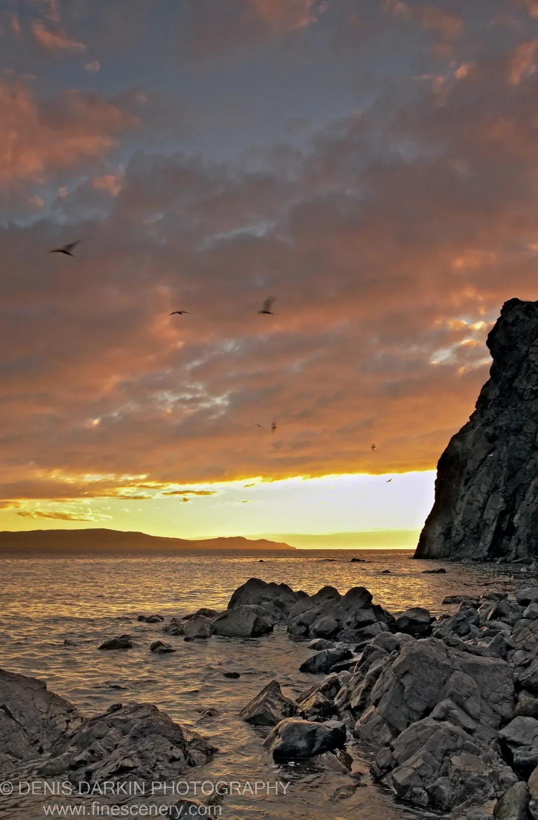 Jagged rocks and a dark cliff face rise above shimmering calm water at dawn on the Russian Far East coast.