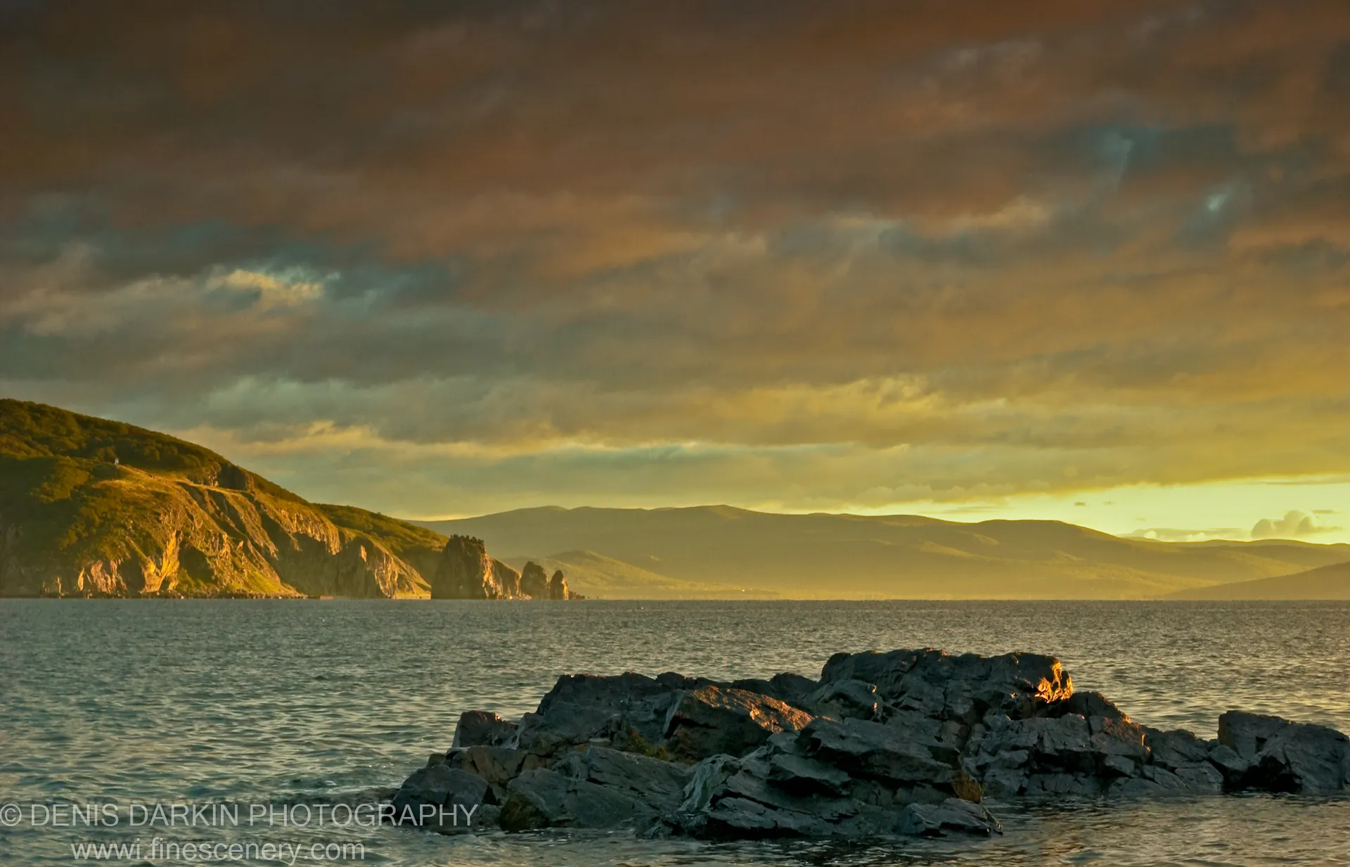 Golden dawn light washes layered rocks and sea cliffs along a coastal bay on the Russian Far East coast.