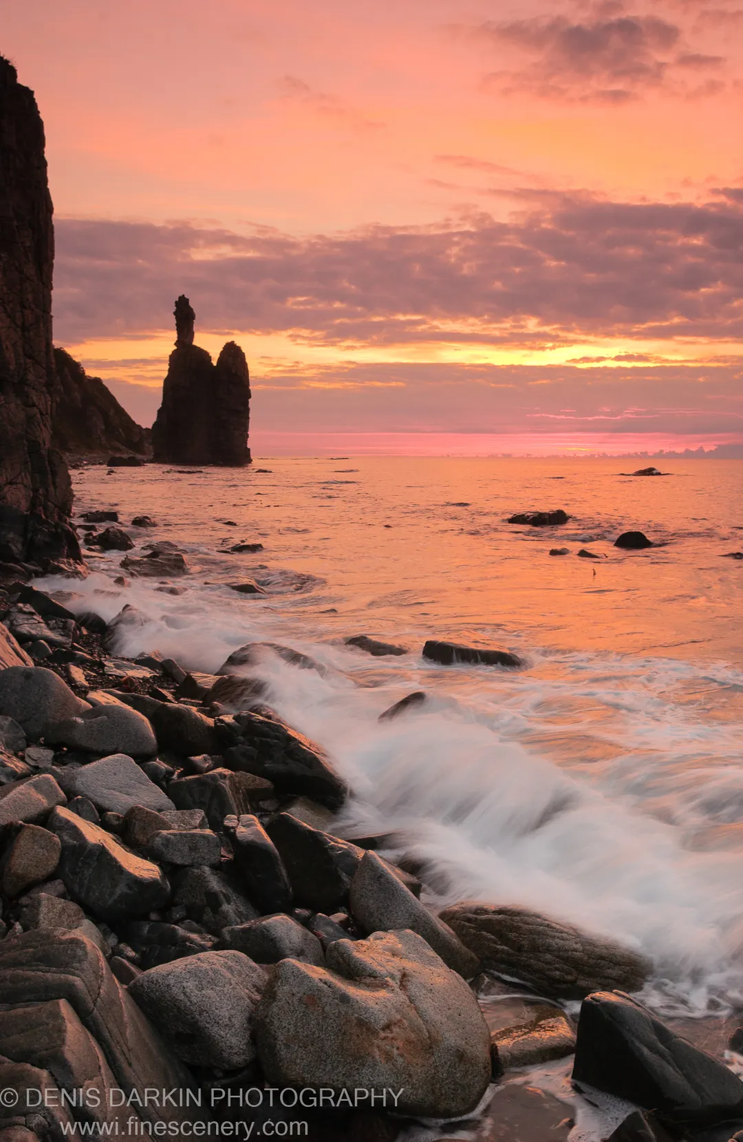 A sea stack stands in silhouette against an orange dawn sky at Plastun, white waves breaking over dark foreground boulders.