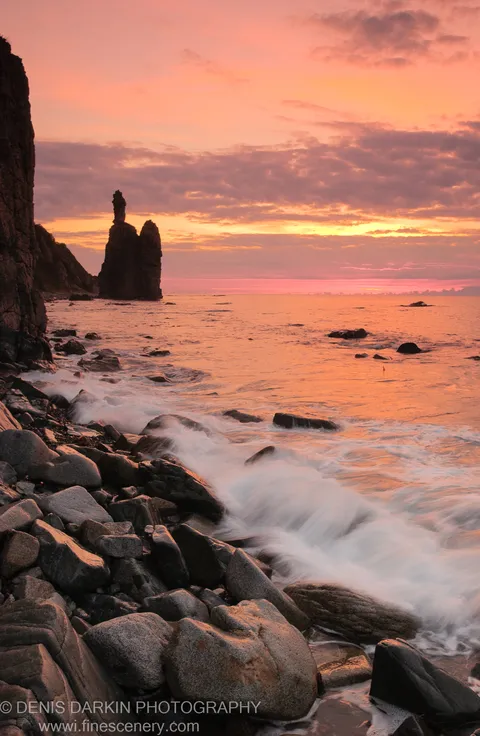 A sea stack stands in silhouette against an orange dawn sky at Plastun, white waves breaking over dark foreground boulders.