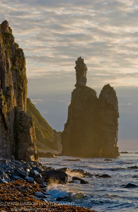 Backlit sea stacks and coastal cliffs stand against a cloud-filled dawn sky near Dalnegorsk, dark rocks and kelp in the foreground where waves break.