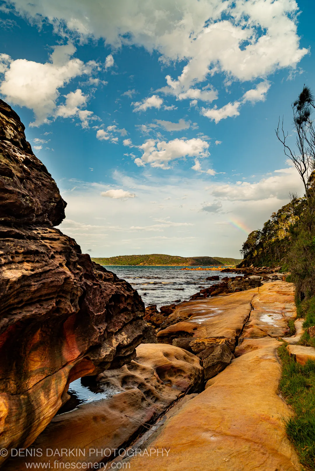 Rock platform and sandstone cliffs above a calm bay at Huskisson, with a faint rainbow over the distant headland. December 2018.