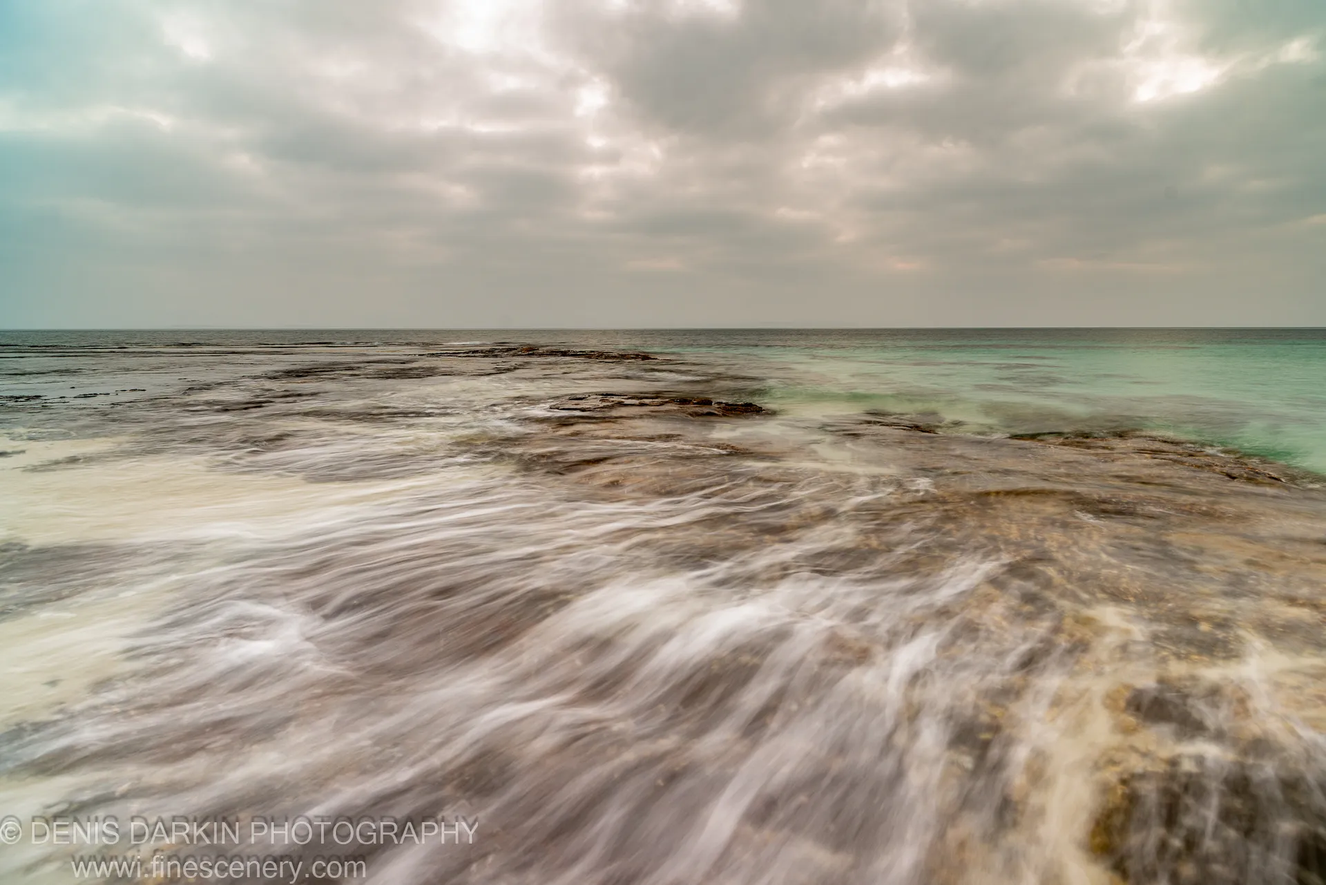 Long-exposure rock platform before sunrise at Huskisson, New South Wales, with motion-blurred surf and patches of turquoise water against an overcast horizon.