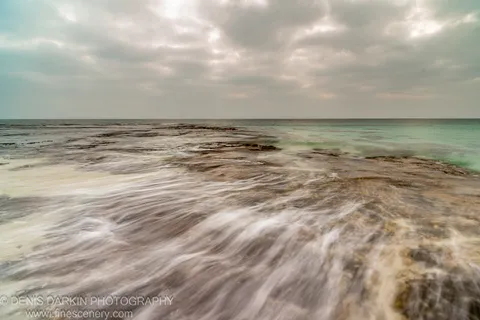 Long-exposure rock platform before sunrise at Huskisson, New South Wales, with motion-blurred surf and patches of turquoise water against an overcast horizon.