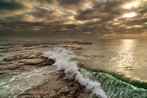 Breaking wave spreading white foam across a flat rock reef at Huskisson at dawn, with dark clouds and shafts of light over the horizon.