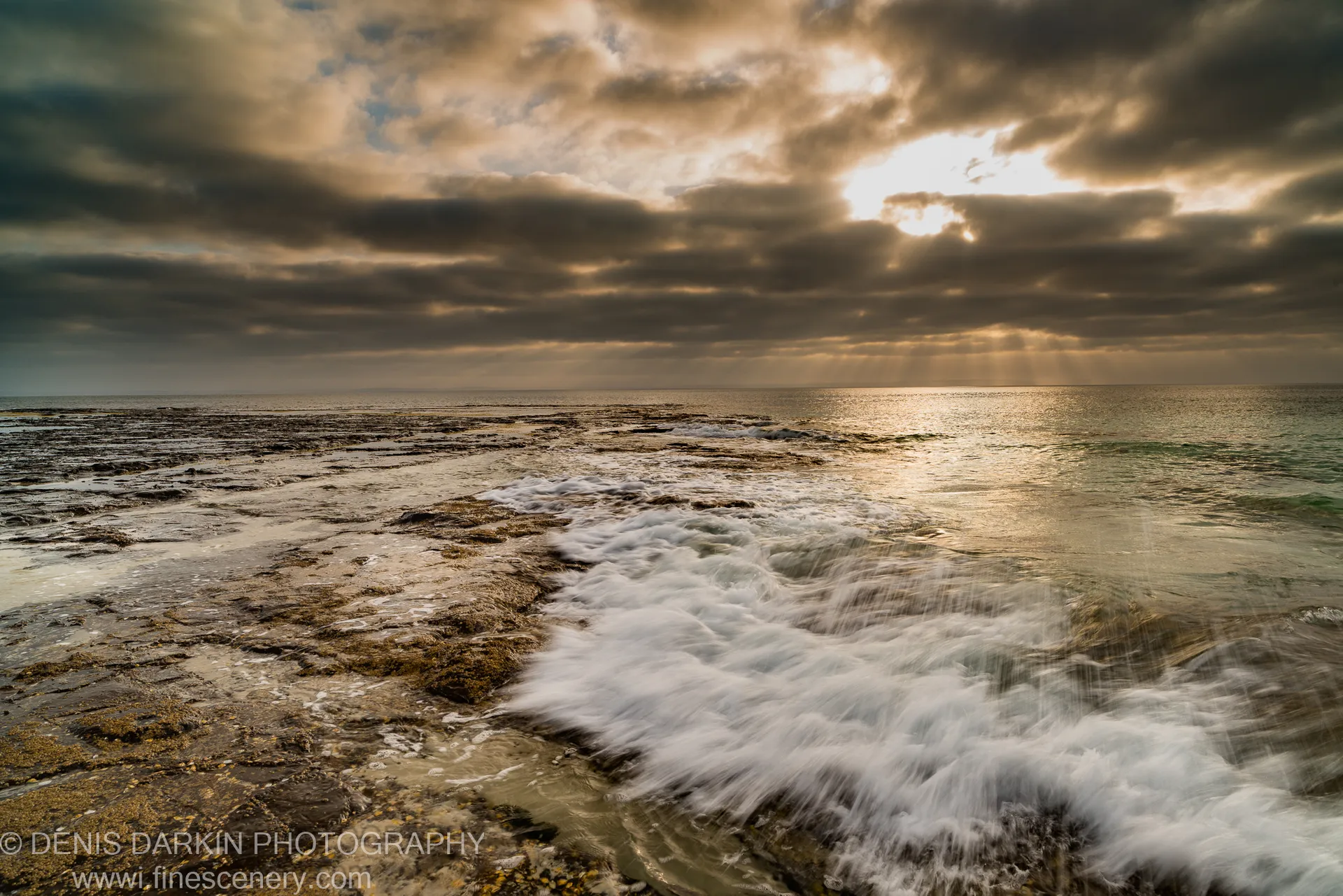 Crepuscular light rays over a tidal shelf at Huskisson at dawn, with white water washing across flat rock and heavy cloud overhead, New South Wales.