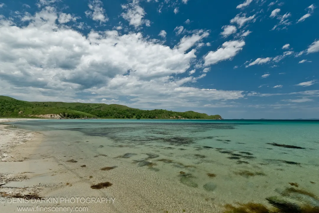 A sandy shoreline transitions into clear turquoise water at Gamov Peninsula, rolling green hills curving around the bay under scattered white cumulus clouds.