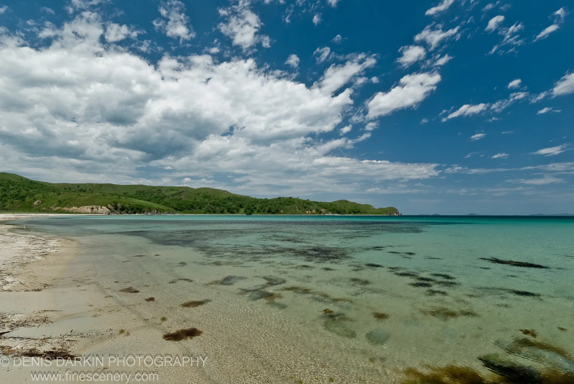A sandy shoreline transitions into clear turquoise water at Gamov Peninsula, rolling green hills curving around the bay under scattered white cumulus clouds.