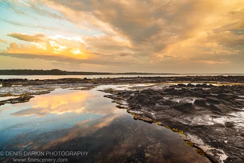 A still tidal pool reflects warm sunrise clouds above a dark basalt shelf on the New South Wales coast.