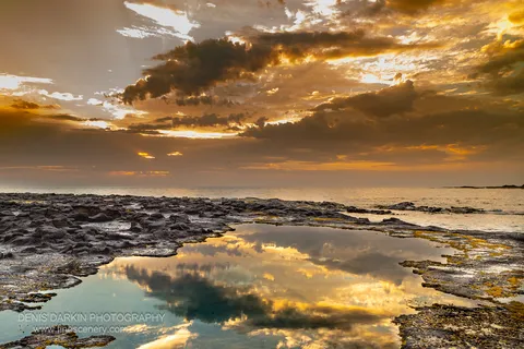 Warm amber light across a tidal flat at dawn, a foreground rockpool reflecting cloud formations and sunbeams, New South Wales coast.